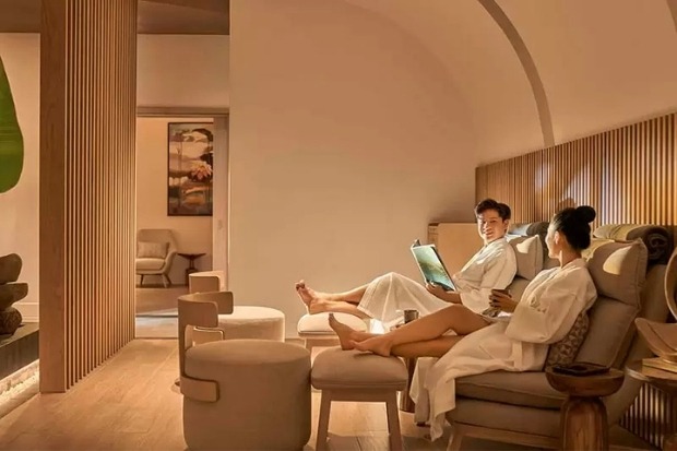 Một cặp đôi mặc áo choàng tắm trắng đang thư giãn trên ghế sofa tại khu vực chờ sang trọng của DuBai Luxury, với không gian được thiết kế hiện đại và ánh sáng ấm áp.