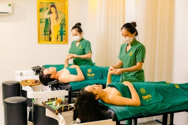 Hai khách hàng, một nam và một nữ, đang nằm thư giãn trên giường trị liệu được phủ khăn màu xanh lá cây đậm và được hai kỹ thuật viên mặc đồng phục xanh, đeo khẩu trang, thực hiện massage tay và vai tại Shinrin-Yoku Spa.